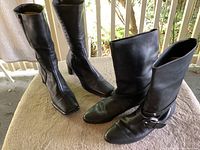 Two pairs of black women's leather boots on display, showing both pairs side by side on a beige surface with railing background.