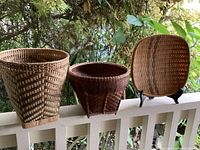 Three vintage bamboo and rattan baskets displayed outdoors on a railing showing different shapes and weaving styles.