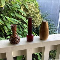 Three rare vintage bud vases displayed outdoors showing diverse materials and shapes: one light wood Myrtle Wood vase, one dark perforated Banksia Nut vase, and one red glass bud vase.