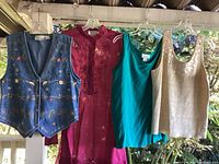 Four vintage women’s tops and vest on hangers, shown outdoors, including an embroidered denim vest, magenta tunic, teal green tank top, and beige sparkly tank.