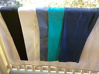 Five pairs of women's slacks displayed side by side, from black to white, steel blue, turquoise, and navy colors.