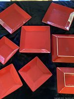 Eight square cherry red ceramic plates and bowls displayed on a dark cloth background. Four plates and four bowls with a smooth, glossy finish and clean edges, no chips or cracks visible.