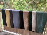 Nine pairs of folded women's pants of various colors displayed outdoors on a railing, showcasing colors including black, beige, gray, taupe, dark green, cream, light blue, and navy.