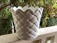 Image showing vintage white ceramic planter with basket weave texture and scalloped rim on an outdoor ledge with greenery background.