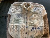 Front view of folded cream-colored wool sweater showing floral knit design and button front.