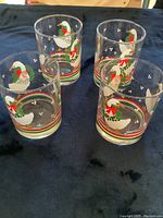 Four clear glasses decorated with white geese wearing Christmas ribbons and holly, displayed on a dark cloth.