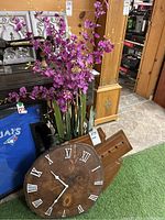 Photo showing wooden wall clock with white Roman numerals and hands in front of faux floral arrangement and display rack