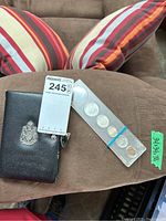 Overview of the coin set in plastic case with black coin case beside it, set displayed on brown cloth surface with striped cushions in background.