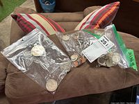 Three plastic bags filled with mixed coins from various countries placed on a brown cushion with striped pillows in the background.