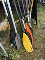 Paddles standing against a yellow rack outdoors, showing aluminum shafts and various colored blades including black and yellow.