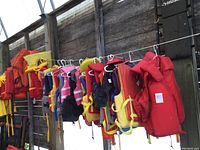 Wide view of various life jackets hanging, colors include red, yellow, black, blue. Different sizes visible from adult to kid.