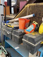 Four black textured resin tackle boxes with yellow clasps on a blue metal shelf alongside orange life jackets, a small lure box, a white hand pump tube, and a yellow flotation device.