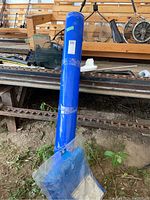 Full view of the blue vinyl roll wrapped in plastic standing vertically outdoors in a workshop area, tarp partially visible at bottom.