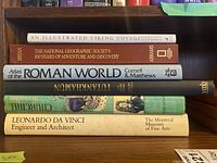 Five hardcover coffee table books stacked and showing titles: An Illustrated Viking Voyage, National Geographic Society 100 Years of Adventure and Discovery, Atlas of the Roman World, Tutankhamun, Churchill Irish Island Farm, Leonardo da Vinci Engineer and Architect.