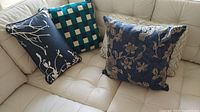 Five decorative pillows arranged on a beige couch; includes a dark blue rectangular pillow with white branches and foliage, green and blue checkered pattern pillow, and dark blue floral embroidered pillow.