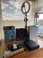 Group shot of all items showing disc drive, ring lamp box, energy lamp box, fan heater, floor lamp, cords and foam tube on table near window.