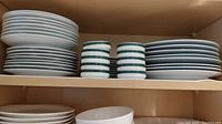 Stacks of white plates and bowls with green and gold rims on a shelf, showing multiple sets of dinner, salad, and bread plates with bowls in the middle, all neatly stacked.