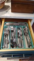 Top view of organized flatware in a drawer with separated compartments showing various forks, knives, spoons, and serving pieces, all in polished stainless steel with decorative handles.