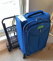Blue TravelPro roller board suitcase standing upright with collapsible luggage dolly behind it near a window.