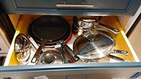 Cookware including stainless steel pots and lids, red and black non-stick frying pans, and a metal teapot on a stovetop.