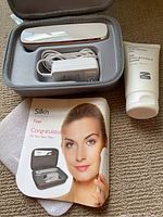 Top-down view of Silk’n Titan device, charger, hyaluronic gel, and user manual in carrying case on carpet.