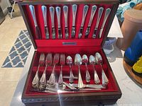 Full view of open wooden display case with silver plated flatware set arranged in slots with red felt lining. Contains knives, forks, spoons, and serving pieces.