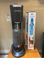 Front view of black Sodastream machine with clear carbonation bottle attached and two Sodastream CO2 cylinders boxed next to it