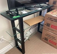 Front angle view of media stand showing the black metal frame and three glass shelves with items on shelves and boxes nearby.