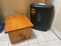 Photo shows wooden stool and black paper shredder side by side on tiled floor.