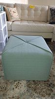 Full front view of the light green square ottoman in front of a white couch on a patterned rug.