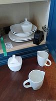 White Thomson pottery creamers, sugar bowl with lid, plates stacked in shelf, and black coasters next to them.