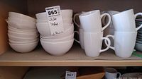 Shelves with stacks of white bowls and white mugs. Stacks of 16 each, simple plain white design.
