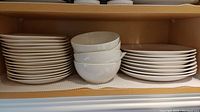Stack of 16 dinner plates, 16 salad plates, and 4 bowls on a shelf, showing overall quantity and condition.
