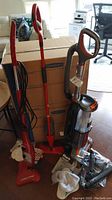 Three cleaning appliances grouped beside cardboard boxes: red H2O Mop Pro X steam cleaner, red mop, and Shark Rocket upright vacuum.