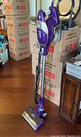 Full view of the Shark Rocket Ultra Light vacuum in purple and silver upright on the floor with power cord wrapped around and some boxes behind it.