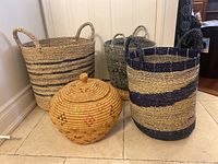 Four woven baskets placed on tiled floor including three tall handled baskets with blue and beige patterns plus one lidded round basket in front