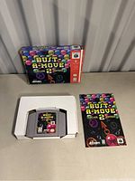 Photo showing N64 game cartridge for Bust-A-Move Arcade 2 Edition with original box and instruction booklet displayed on a surface.
