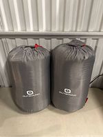 Two gray Outbound sleeping bags in compression sacks on the floor against a metal wall.