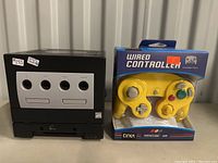 Front view of black Nintendo GameCube console on top of the attached GameCube Player add-on, showing four controller ports and ATI logo; yellow Cirka wired controller in unopened box to the right.