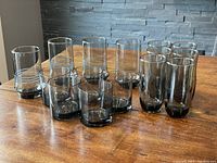 Full set of 15 smokey retro drinking glasses arranged on wood table with grey stone background.