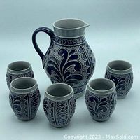 Full set photo showing one pitcher and five matching cups in blue and grey salt glaze with decorative raised patterns.