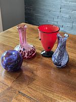 Overview of all four colourful signed studio glass pieces shown on wooden surface: a pinkish slender vase, a bright red art deco style vase with black base, a purple swirl apple paperweight, and a smaller purple swirl bud vase.