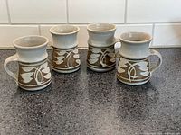 Photo showing four ceramic mugs with gray upper portions and brown lower portions decorated with white leaf and branch designs, arranged on a dark countertop.