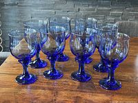 Eight Mediterranean blue cobalt stemmed glasses arranged on a wooden table showing the full set and their shape.