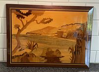 Full view of wood inlay marquetry artwork showing coastal scene with tree, water, buildings, rocks and framed border
