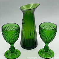 Set of emerald green glass carafe and two matching goblets with diamond point detailing shown upright on white background.