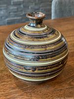 Front and side view of a round striped ceramic vase with brown, black, cream, and gold horizontal bands on a wooden table.