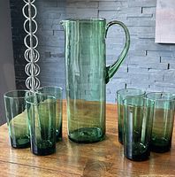 Green glass pitcher with six matching vintage green glasses arranged on a wooden surface in front of a stone wall.