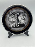 Full front view of the decorative plate on display stand showing the intricate black and white sunshine and female profile design with gold rim.