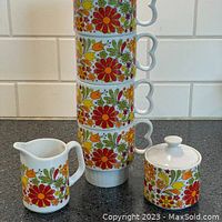Stacked view of four colorful floral mugs with creamer and covered sugar bowl on the side.